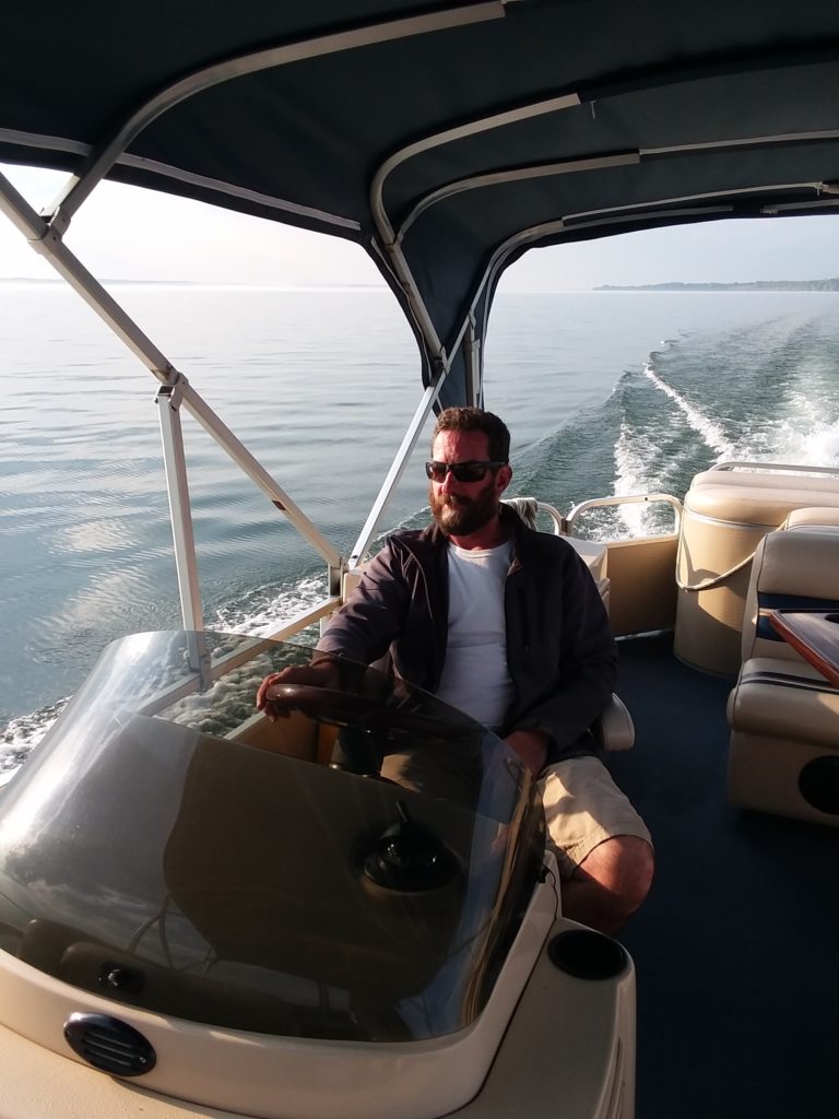 Captain Matt Bianconi, wearing sunglasses, sits in the captain's chair of a speeding pontoon boat on the lake