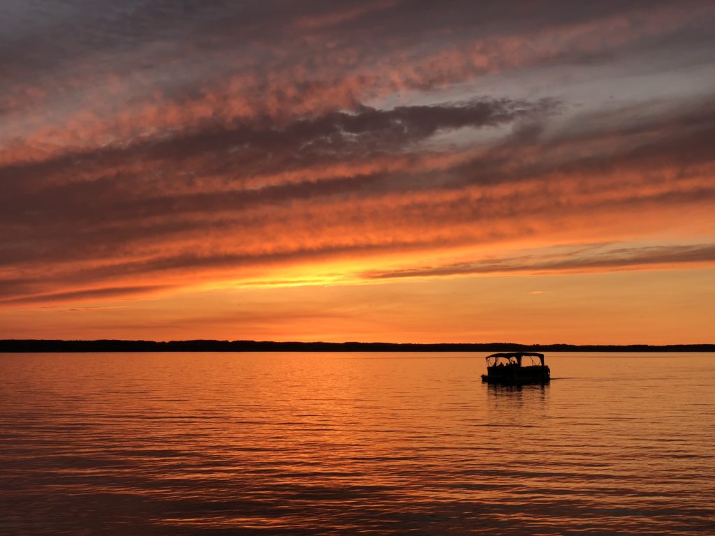 A pontoon boat cruises Cayuga Lake's calm waters under a bright yellow and orange sunset sky.
